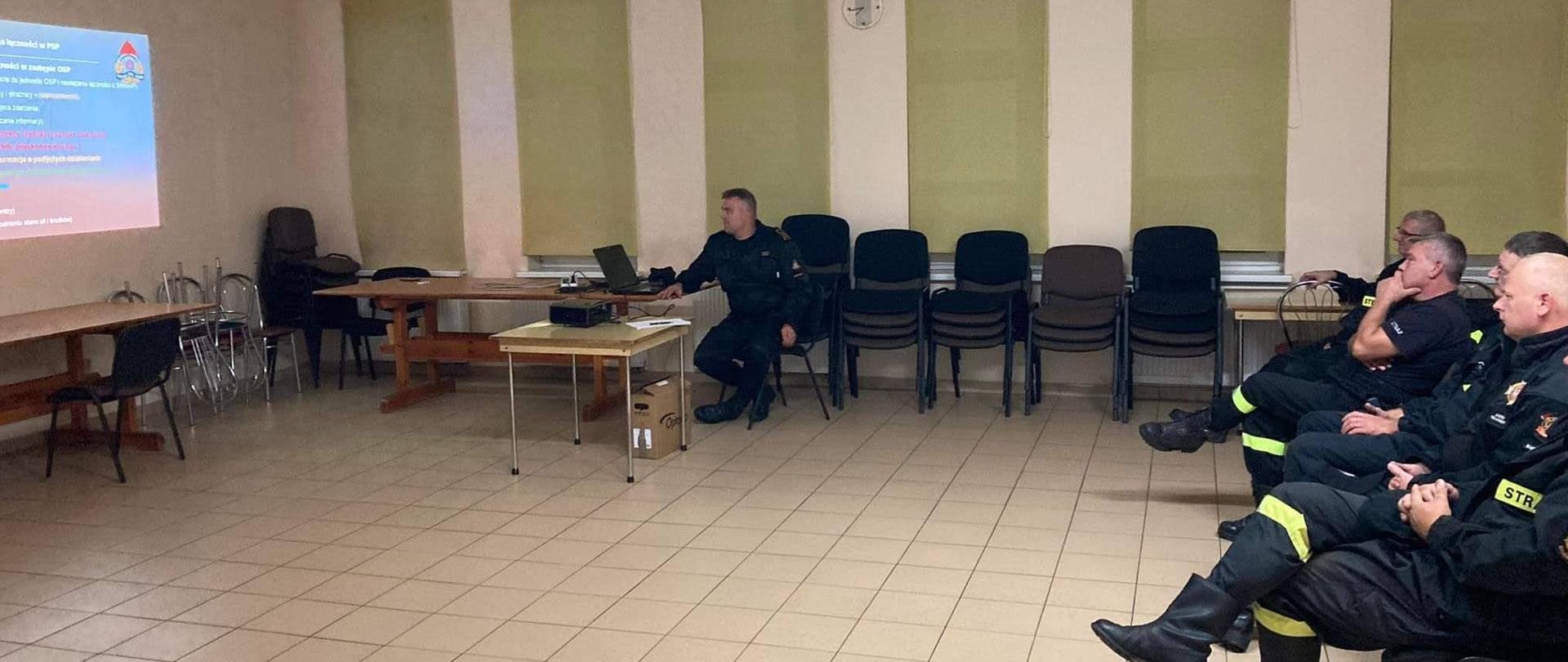 W sali na terenie wyznaczonej remizy OSP odbywa się szkolenie z zakresu organizacji łączności radiowej dedykowane dla druhów jednostek OSP z terenu powiatu krotoszyńskiego. Druhowie zasiadający na krzesłach zapoznają się z materiałem wyświetlanym na prezentacji multimedialnej przygotowanej przez funkcjonariusz prowadzącego szkolenie. Funkcjonariuszem prowadzącym szkolenie jest strażak z krotoszyńskiej komendy który omawia słuchaczom prezentowany materiał.