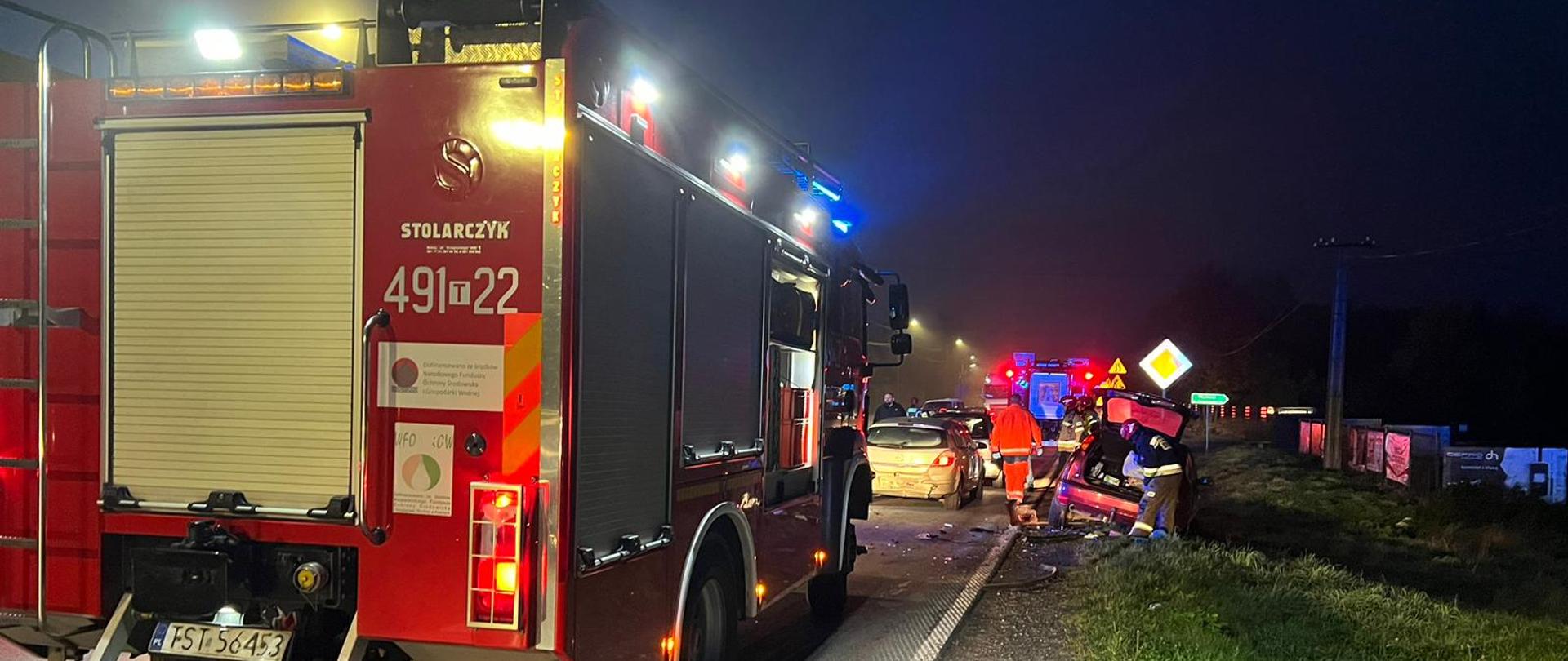 Samochody pożarnicze na sygnałach świetlnych, na jezdni, w tle rozbity pojazd osobowy