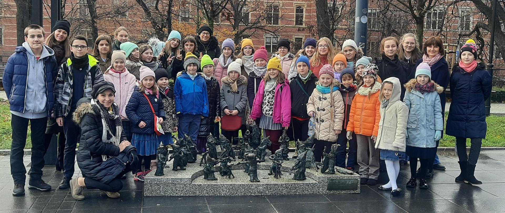 Zdjęcie przedstawiające grupę uczniów i nauczycieli. Grupa stoi na zewnątrz Narodowego Forum Muzyki przy figurach krasnoludkowej orkiestry symfonicznej , w tle budynek wrocławskiego sądu (zbudowany z czerwonej cegły).