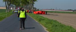 Tragiczny wypadek. Droga z Sarnówki do Łaszczyna. Na pierwszym planie policjanci w kamizelkach odblaskowych z napisem "POLICJA" dokonujący oględzin. W tle miejsce wypadku - widoczny fragment rozbitego auta, wokół którego rozstawione sa czerwone parawany ochronne. Z lewej strony na jezdni stoi radiowóz policyjny i wóz strażacki. W głębi drzewa i zabudowania. 