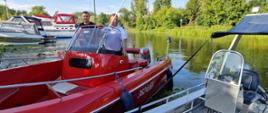 Zdjęcie przedstawia łódź strażacką, na które znajduje się strażak oraz policjant. Łódź stoi przy pomoście na jeziorze. Obok zaparowane łodzie prywatne. 
