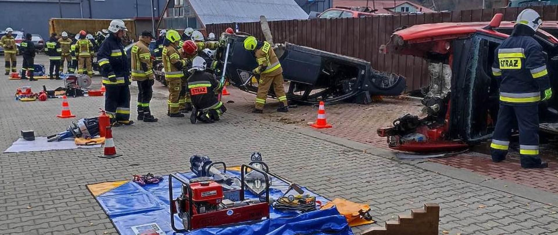 Kolorowe zdjęcie wykonane w jasny dzień, przedstawia utwardzony plac na którym widać kilka grup umundurowanych na bojowo strażaków. Ćwiczą ze sprzętem ratowniczym przy kilku wrakach pojazdów. Sprzęt przygotowany i zgromadzonych na niebieskich matach przed wrakami. W tle widoczne częściowo zabudowania, ogrodzenie i szare drzewa. 