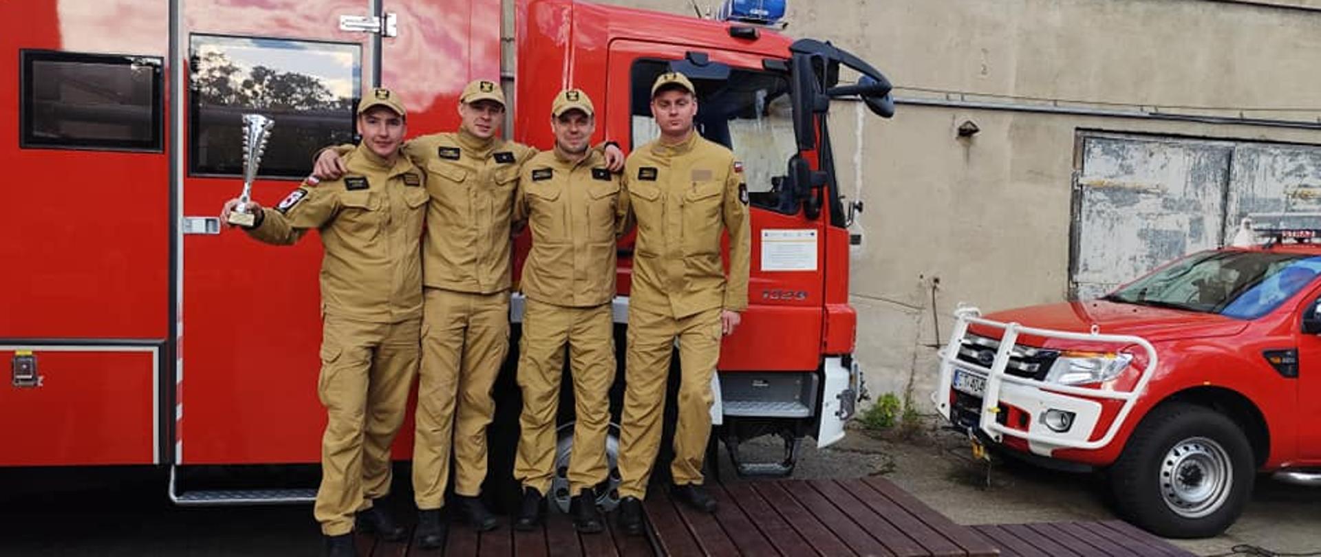 Zdjęcie przedstawia drużynę Komendy w Brodnicy na podium Kujawsko-Pomorskich Zawodów Strażackich w Ratownictwie Wysokościowym. Strażak po lewo unosi w dłoni puchar za zdobycie drugiego miejsca. Za nimi zaparkowano dwa samochody Straży Pożarnej.
