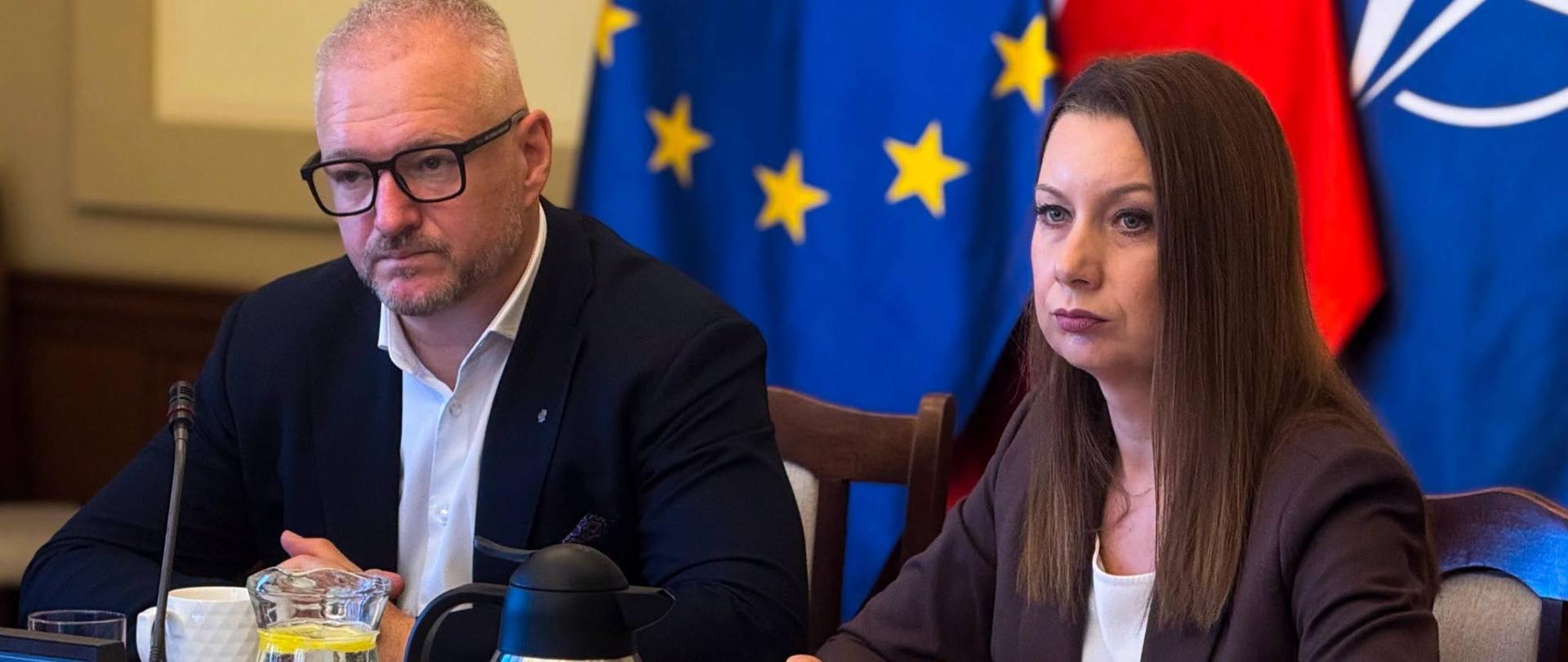 Wojewoda warmińsko-mazurski oraz dyrektor generalny Urzędu Wojewódzkiego siedzą przy stole i uważnie słuchają wypowiedzi uczestnika spotkania. Mają poważne miny. W tle widoczne są flagi Unii Europejskiej, Polski i NATO.
