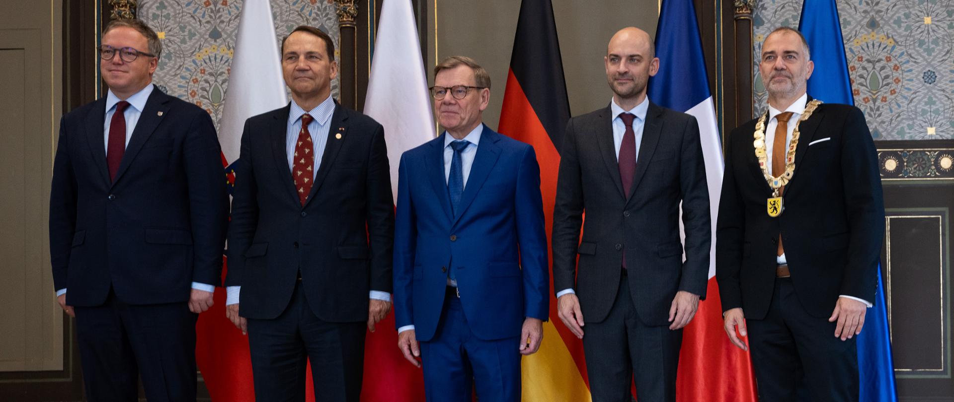The photo shows five men in suits. They are standing and looking straight ahead. Behind them are five flags.