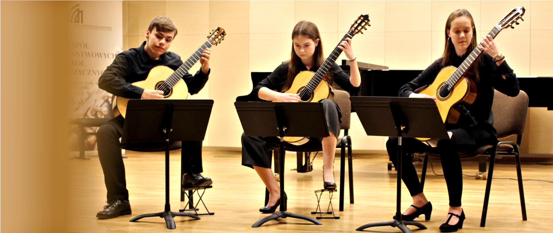 Zdjęcie przestawia trzech uczniów ZPSM grających w sali koncertowej na gitarach