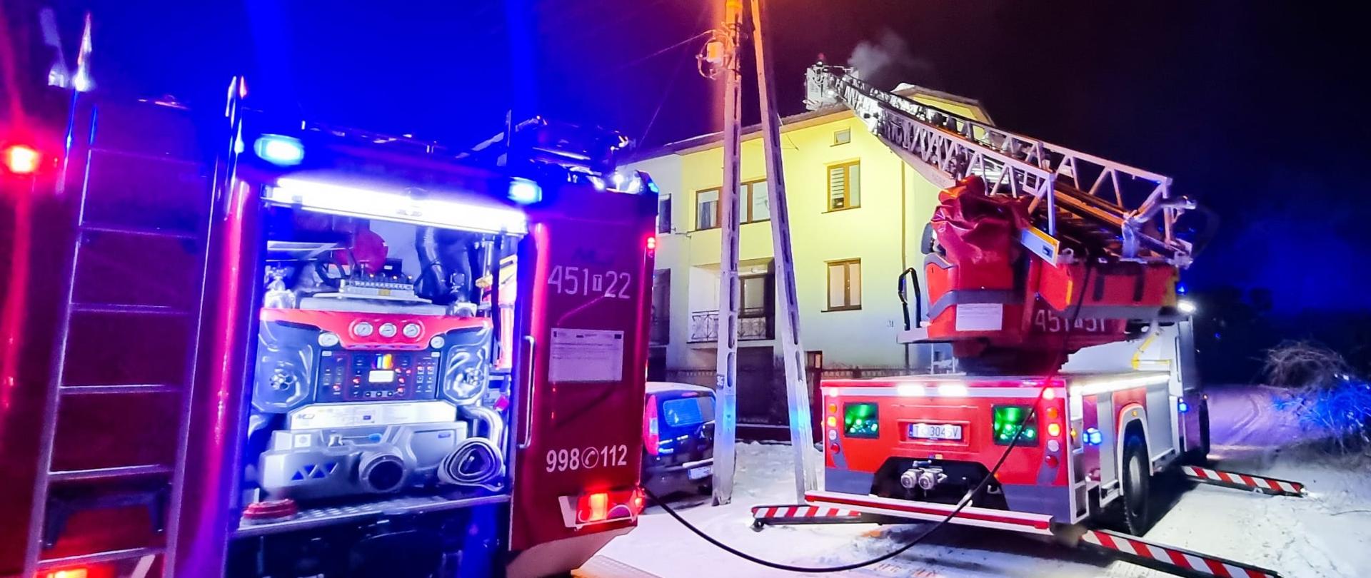 Zdjęcie z pożaru sadzy w przewodzie kominowym. Na pierwszym planie rozstawiona samochód drabina pożarnicza oraz samochód gaśniczy. W oddali strażak w koszu drabiny nad budynkiem mieszkalnym.