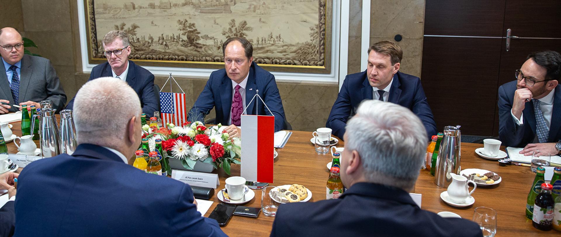 Wicepremier Jacek Sasin siedzi przy stole, obok siedzi inny mężczyzna. Po przeciwnej stronie delegacja amerykańska. Na stole stoją proporczyki i kwiaty. 