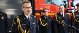 Zastępca Komendanta Powiatowego Państwowej Straży Pożarnej starszy brygadier Andrzej Rafalski przemawia przy mównicy. W tle stoją funkcjonariusze Państwowej Straży Pożarnej. 