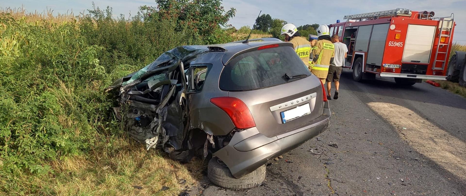 Zdjęcie przedstawia samochód osobowy po wypadku z uszkodzonym lewym bokiem, obok stoją strażacy i osoba postronna. Po prawej stronie znajduje się wóz strażacki