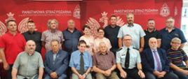 Zdjęcie grupowe osób ustowiomych w dwóch rzędach, pierwszy rząd siedzi na krzesłach, w tle czerwone ścianki reklamowe z napisem Państwowe Straż Pożarna