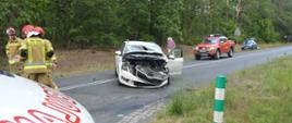 Zderzenie aut na skrzyżowaniu. Na pierwszym planie fragment ambulansu oraz biała skoda octavia z uniesioną pokrywą silnika i uszkodzonym przodem. Na jezdni stoi trzech strażaków w czerwonych hełmach. Z prawej strony skody znajduje się wóz straży pożarnej. W tle - las.