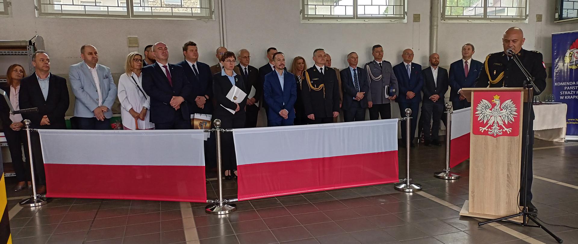 Na trybunie honorowej zaproszeni na Uroczystość goście /Po prawej stronie na mównicą przemawiający Komendant Powiatowy PPS w Krapkowicach w mundurze galowym, bez czapki. uroczystość odbywa się na garażu JRG. 