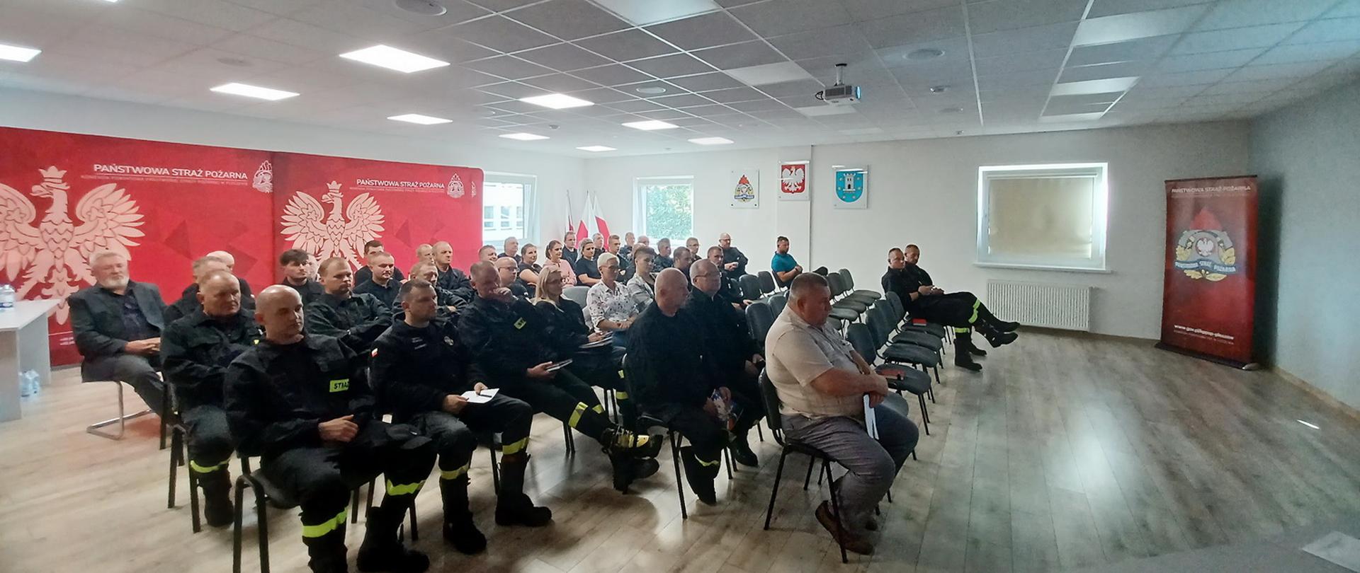 Kilkunastu strażaków OSP siedzi na zaciemnionej sali, część z nich jest w mundurach, pozostali w ubraniach cywilnych