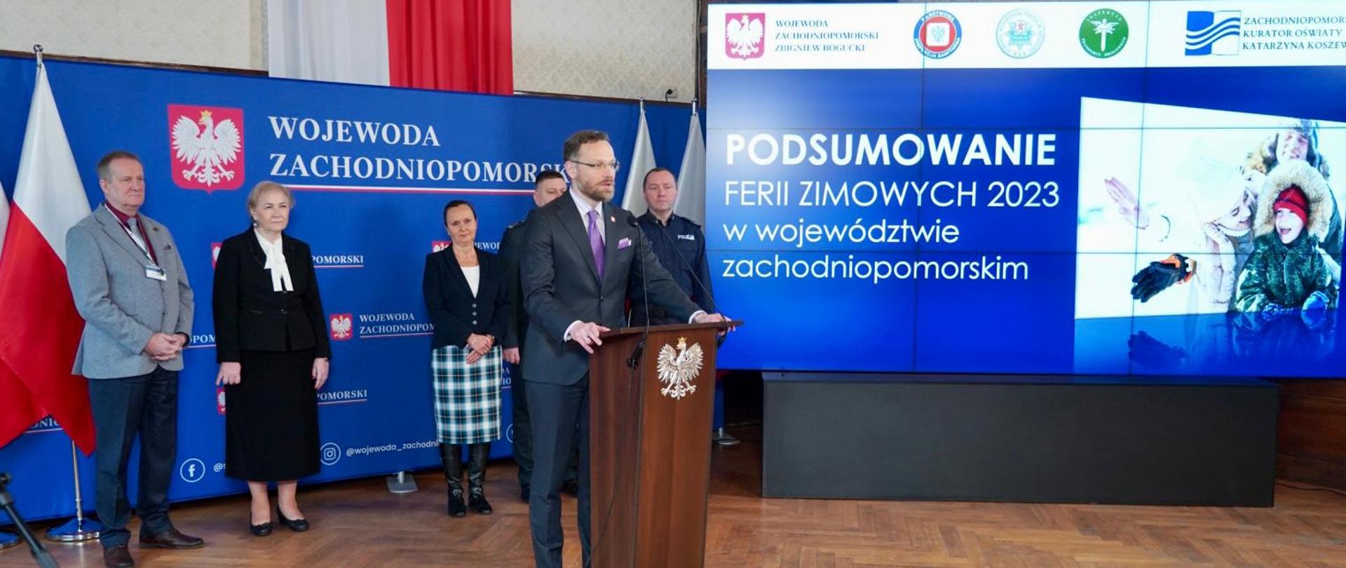 wojewoda Zbigniew Bogucki przy mównicy, za nim szefowie służb, w tle slajd z tytułem konferencji: podsumowanie ferii zimowych 2023 w województwie zachodniopomorskim