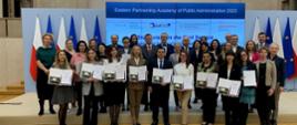 Uczestnicy i organizatorzy wizyt studyjnych w ramach Akademii Administracji Publicznej Partnerstwa Wschodniego 2023 - grupa około 30 osób stoi w dwóch rzędach na tle elektronicznego baneru oraz flag Polski i Unii Europejskiej; w pierwszym rzędzie uczestnicy projektu z pamiątkowymi dyplomami 