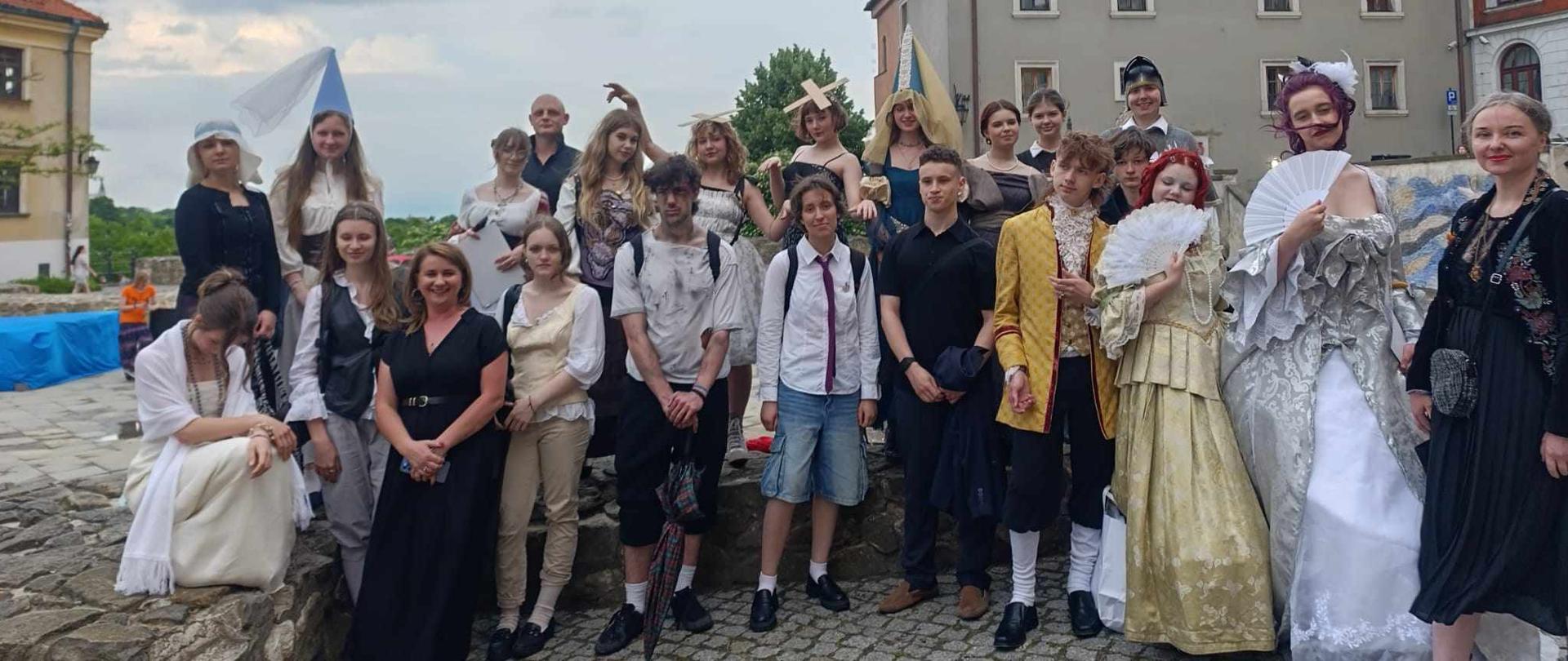 Grupowe zdjęcie wykonane na świeżym powietrzu, na tle zabytkowej zabudowy miejskiej. Na fotografii widzimy dużą grupę młodych osób oraz kilka dorosłych, pozujących razem na brukowanym placu. Większość osób ubrana jest w historyczne stroje z różnych epok – od średniowiecza po XVIII wiek – w tym suknie, peruki, wachlarze, stroje chłopskie i szlacheckie. W tle widać zabytkowe kamienice z czerwonymi dachami oraz fragment nieba z chmurami. Atmosfera zdjęcia jest radosna i teatralna – prawdopodobnie dokumentuje udział grupy w wydarzeniu edukacyjno-historycznym lub inscenizacji, np. w ramach szkolnego projektu lub imprezy kulturalnej. 