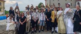 Grupowe zdjęcie wykonane na świeżym powietrzu, na tle zabytkowej zabudowy miejskiej. Na fotografii widzimy dużą grupę młodych osób oraz kilka dorosłych, pozujących razem na brukowanym placu. Większość osób ubrana jest w historyczne stroje z różnych epok – od średniowiecza po XVIII wiek – w tym suknie, peruki, wachlarze, stroje chłopskie i szlacheckie. W tle widać zabytkowe kamienice z czerwonymi dachami oraz fragment nieba z chmurami. Atmosfera zdjęcia jest radosna i teatralna – prawdopodobnie dokumentuje udział grupy w wydarzeniu edukacyjno-historycznym lub inscenizacji, np. w ramach szkolnego projektu lub imprezy kulturalnej. 