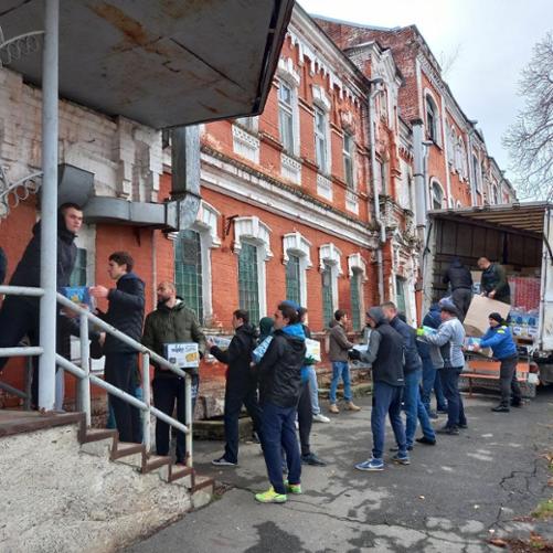 Przed budynkiem grupa mężczyzn rozpakowuje samochód z pomocą humanitarną, przekazując sobie paczki z rąk do rąk