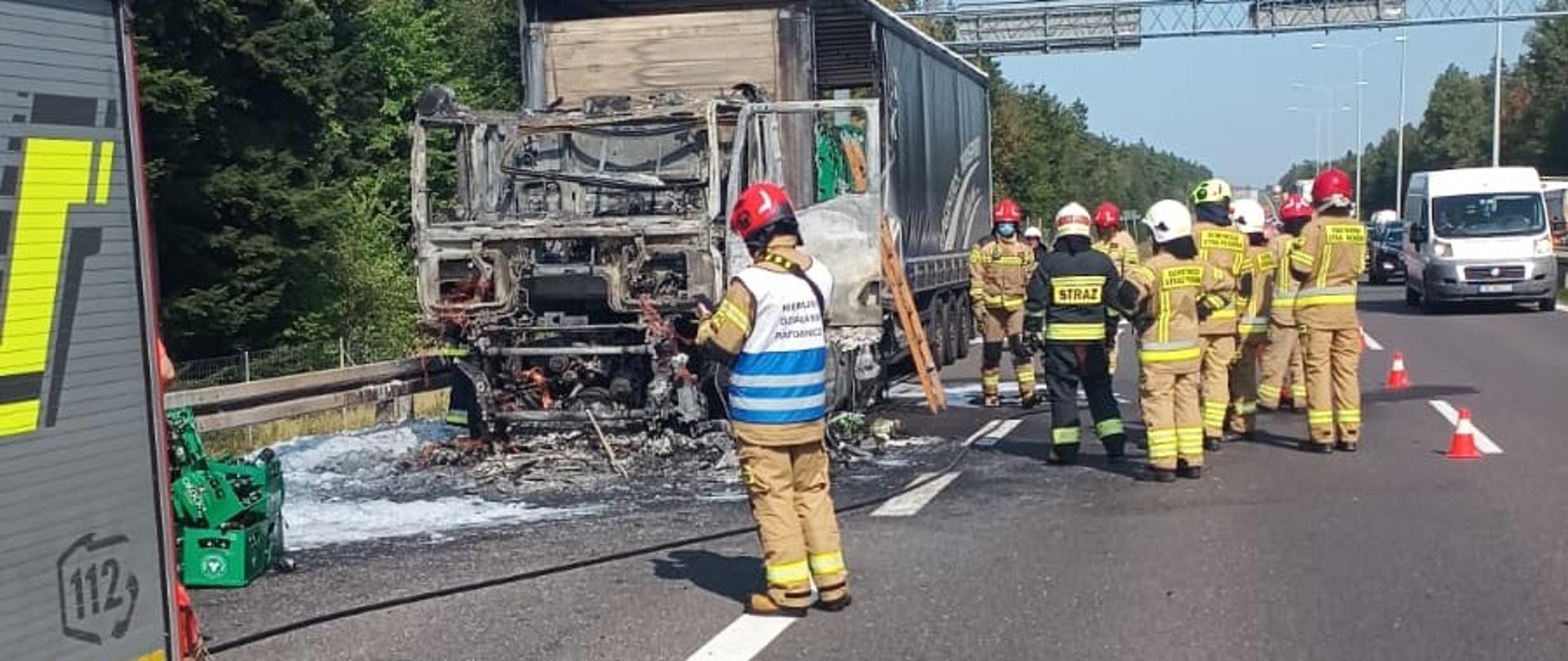 Zdjęcie przedstawia miejsce drogę S7 gdzie doszło do pożaru samochodu ciężarowego. Po lewej stronie stoi spalony ciągnik siodłowy, przy którym na jezdni, w centralnym punkcie zdjęcia znajdują się ratownicy.