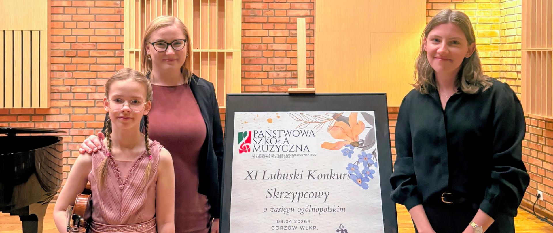 Treść wiadomości Gemini
Dziewczynka ze skrzypcami oraz dwie kobiety stojące w sali obok sztalugi z plakatem XI Lubuskiego Konkursu Skrzypcowego, w tle ściana z cegły, drzwi i okno