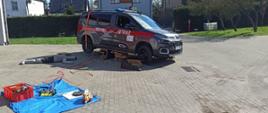 Zdjęcie przedstawia samochód strażacki. Auto małe, dostawcze i oznakowane. Pod autem osoba poszkodowana, manekin. Jest dzień, pogodnie i słonecznie.