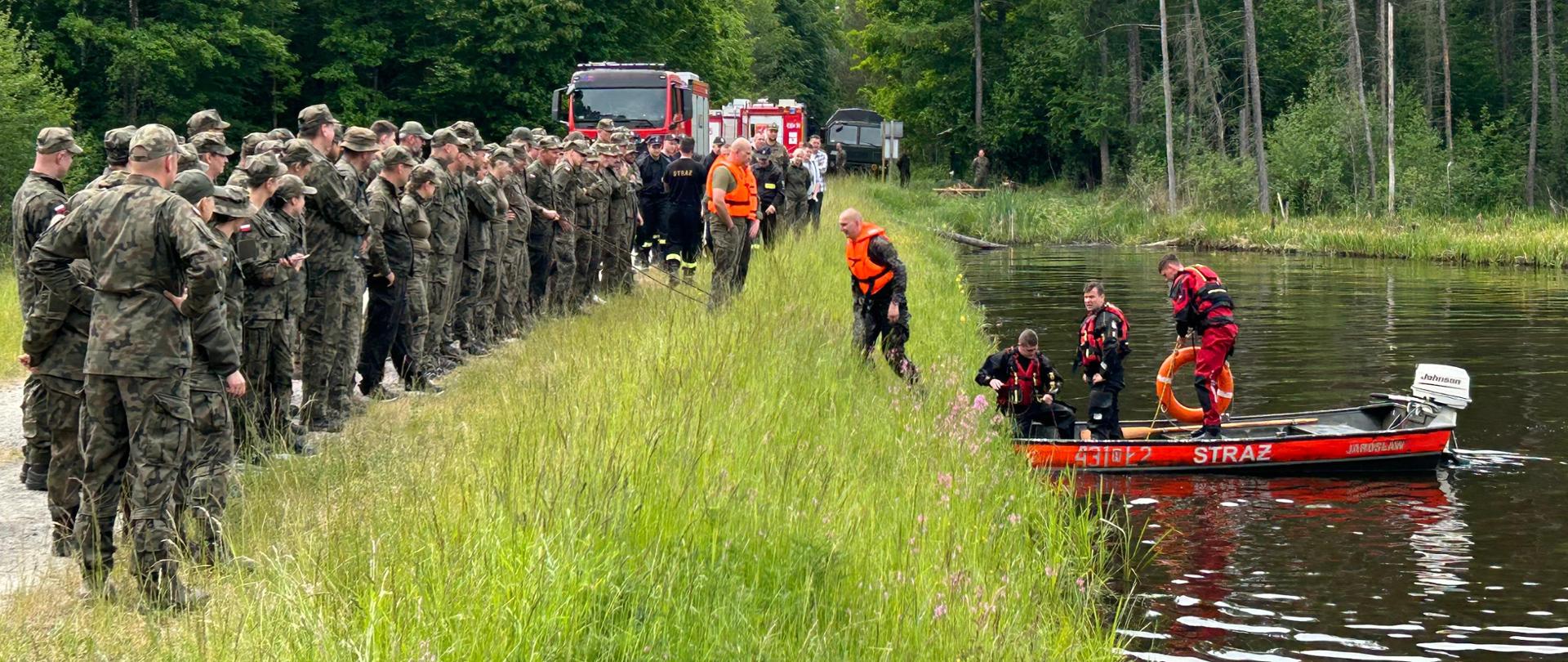 Na zdjęciu widzimy strażaków ubranych w skafandry wypornościowe którzy wodują łódź na zbiorniku wodnym. Obok niego znajduje się torba medyczna. Na brzegu, w dwuszeregu zgromadzeni są żołnierze, którzy obserwują działania strażaków. W tle widoczny jest las.