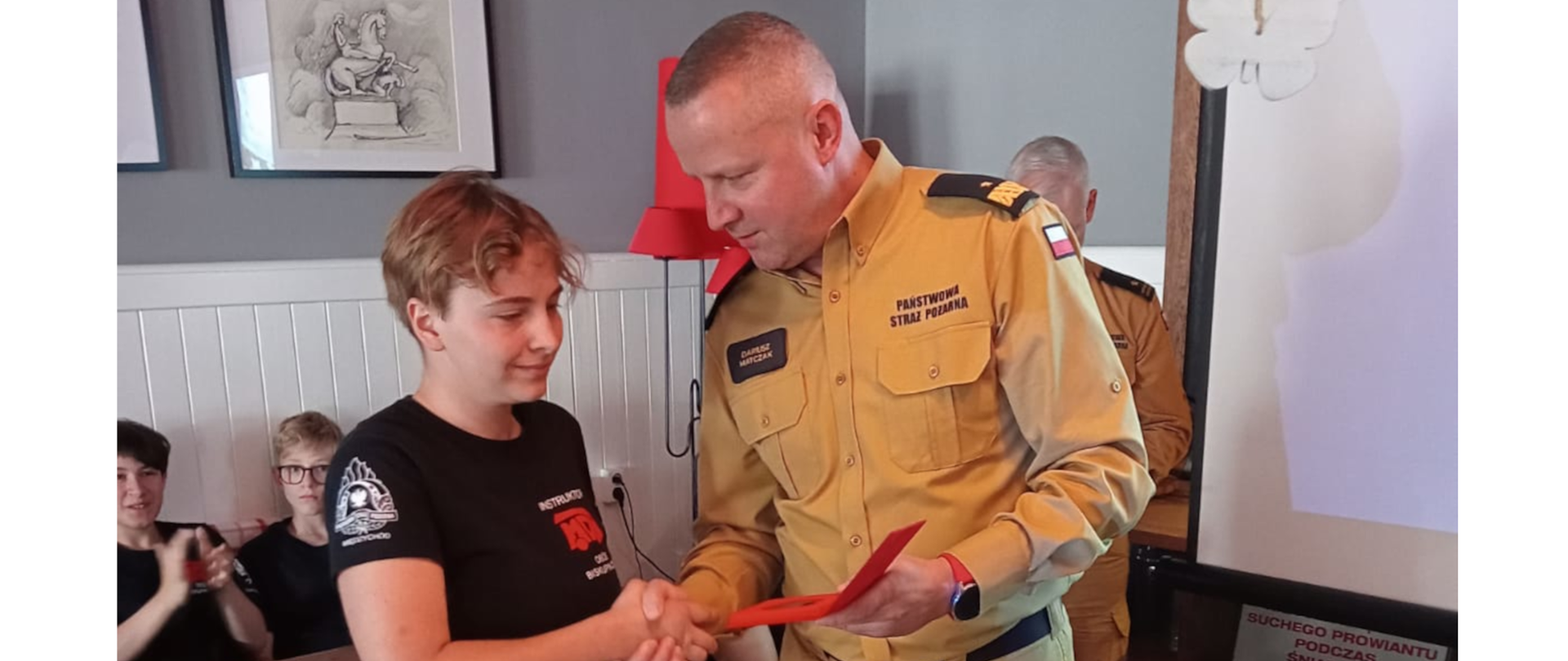 strażak ściska dłoń chłopcu , wręcza mu medal, w tle dzieci przy stołach