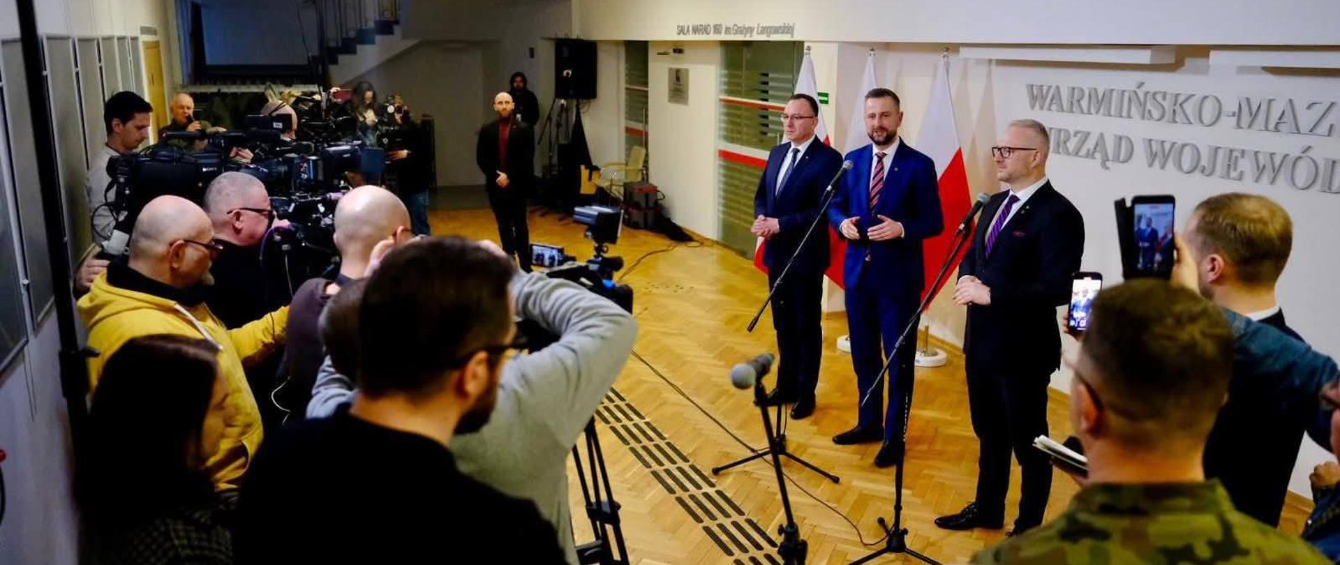 tłum ludzi z mikrofonami i kamerami patrzą w kierunku 3 mężczyzn którzy stoją na tle ściany Warmińsko-Mazurskiego Urzędu Wojewódzkiego. Mówią przez mikrofon