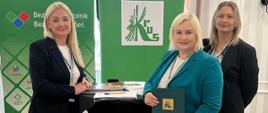 Trzy kobiety stojące przy wysokim stoliku na sali konferencyjnej. Za nimi znajdują się zielone banery z nazwą KRUS. Kobiety są uśmiechnięte i ubrane elegancko. Jedna z nich trzyma teczkę z logo KRUS.
