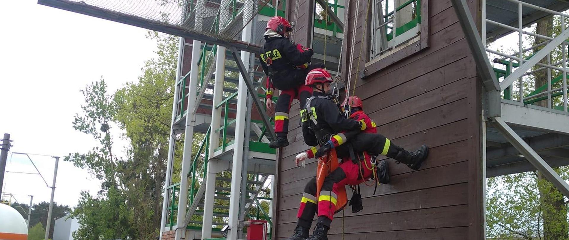 ściana wspinalni na ścianie dwóch ratowników w czerwonych skafandrach i dwóch w czarnych ubraniach koszarowych