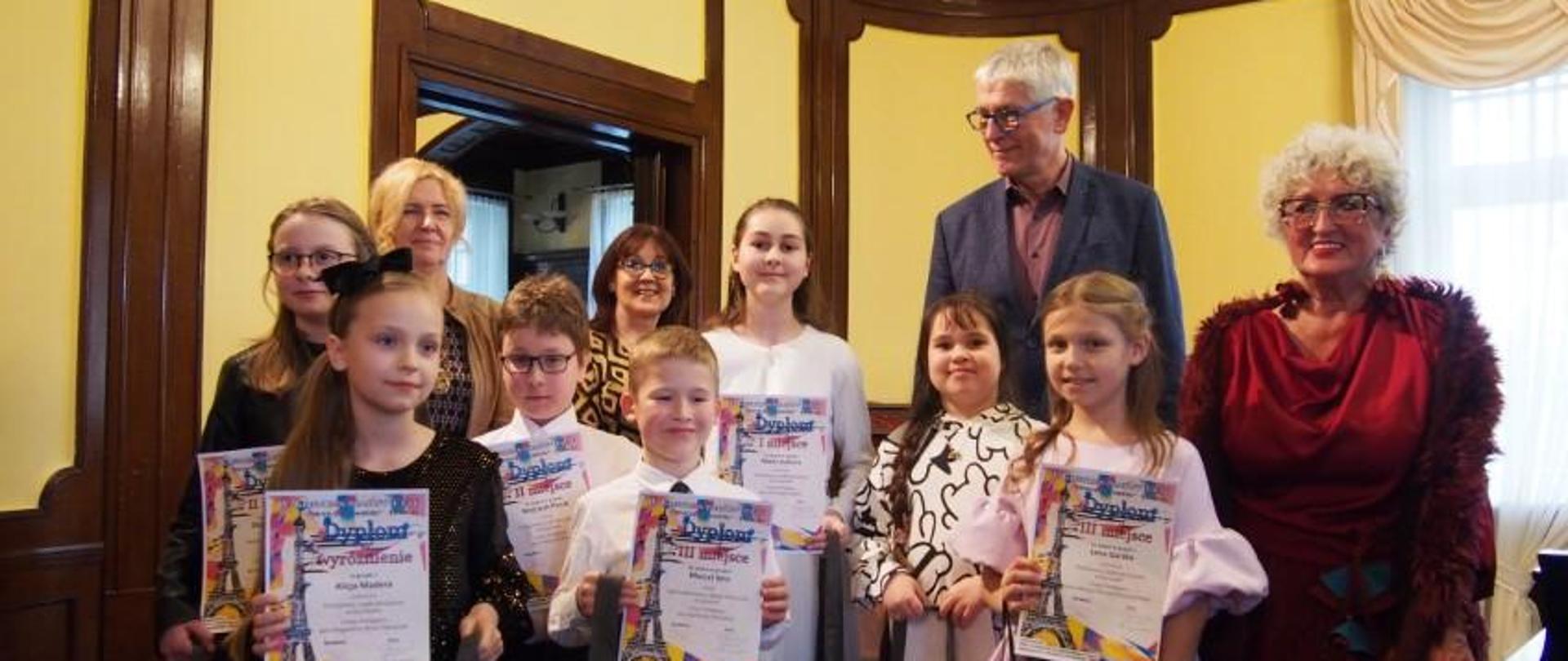 Zdjęcie kolorowe: ellgancko ubrane dzieci - laureaci - siedmioro uczestników prezentujących swoje dyplomy; za nimi stoją członkowie jury i pani dyrektor szkoły muzycznej ze Słupska.