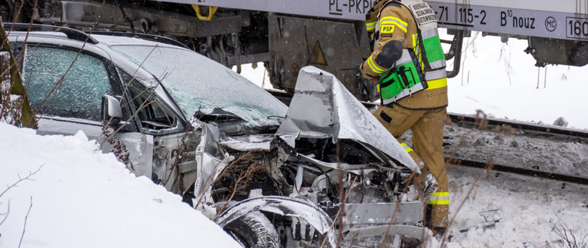 Zdjęcie przedstawia zniszczony samochód koloru srebrnego po zderzeniu z pociągiem w miejscowości Poronin koło Zakopanego. W tle widać strażaka oraz pociąg. Na zdjęciu widać również śnieg.