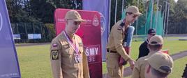 Uczestnicy wydarzenia w umundurowaniu podczas ceremonii dekoracji na podium. Jeden z zawodników stoi na stopniu oznaczonym numerem 2, a pozostali gromadzą się obok podium. W tle znajdują się banery promocyjne, drzewa i teren sportowy.