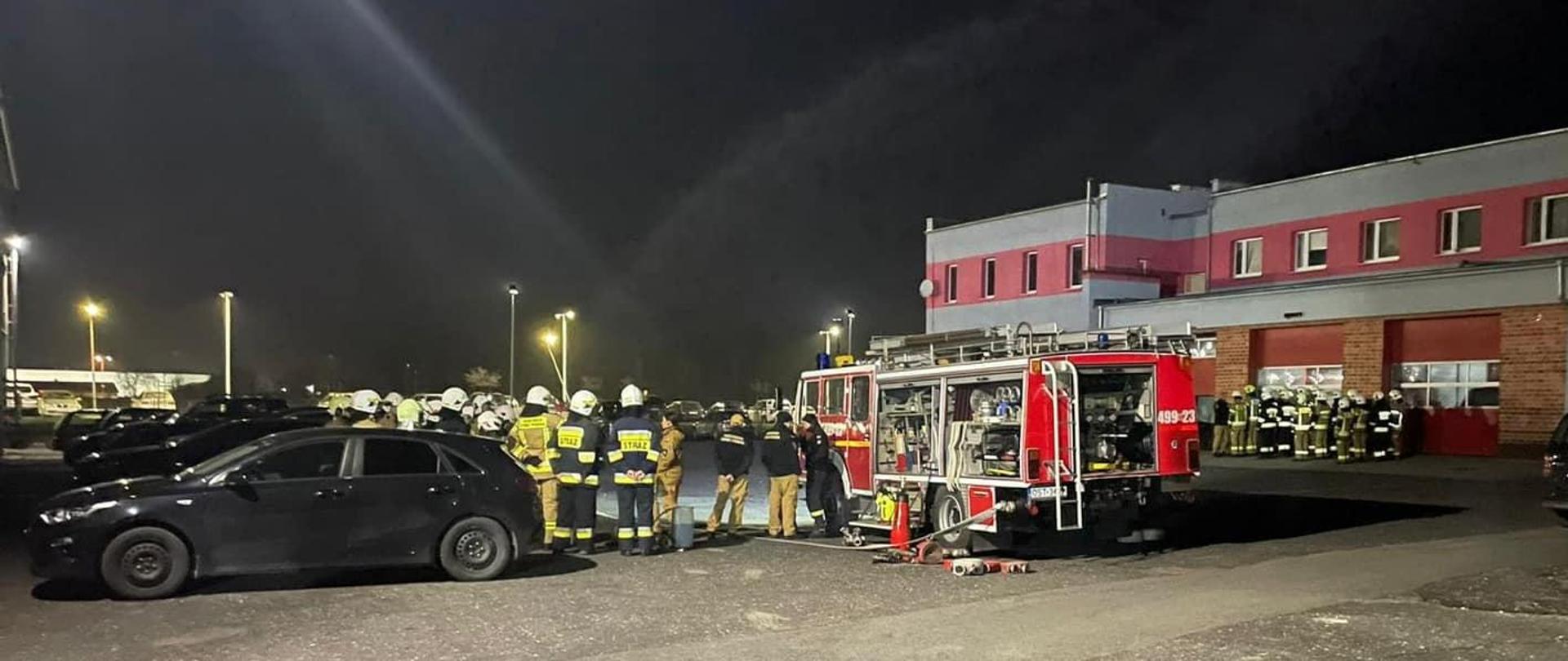 Na zdjęciu znajdują się strażacy ubrani w ubrania specjalne, ustawieni przy samochodach strażackich i osobowych. W tle widać budynki komendy oraz samochód strażacki z wyciągniętym sprzętem