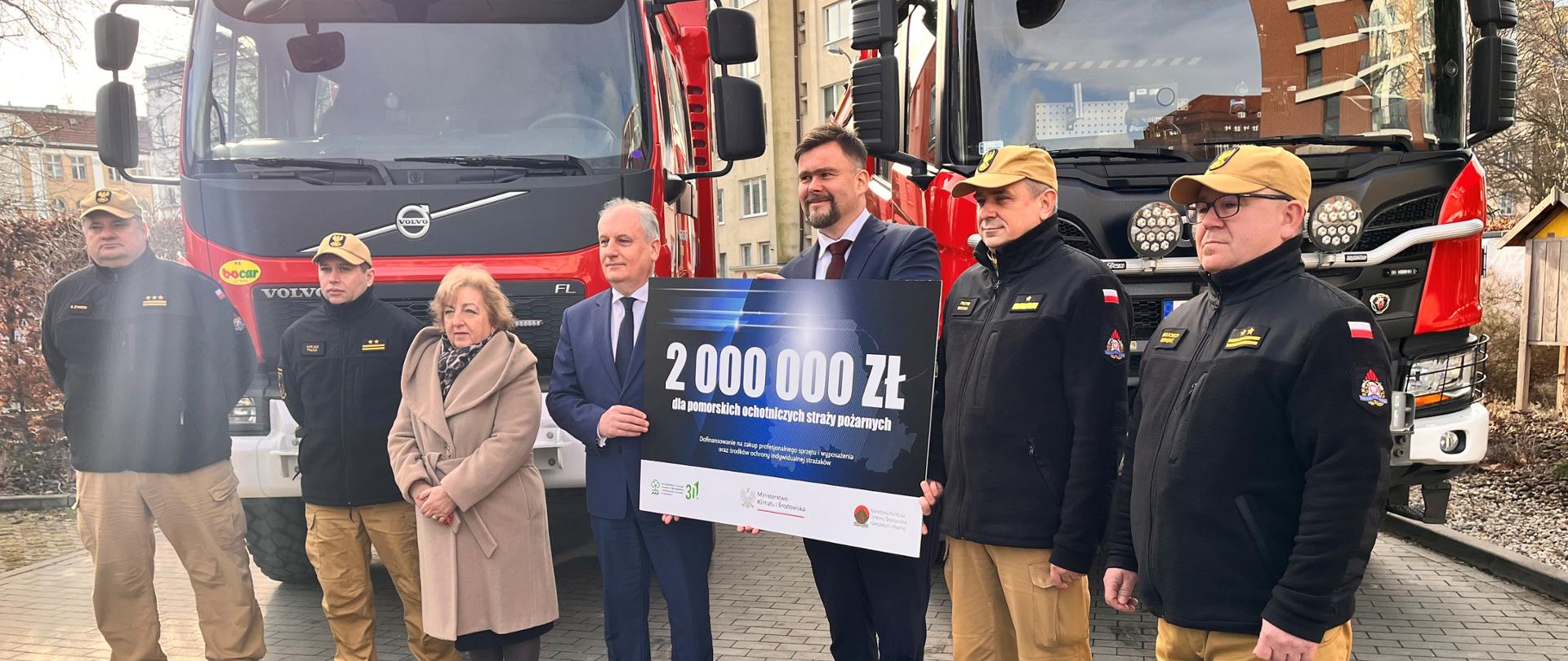 Strażacy oraz osoby cywilne mężczyźni oraz jedna kobieta stoją przed samochodami gaśniczymi dwóch mężczyzn trzyma tablicę z liczbą dwa miliony złotych. 