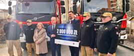 Strażacy oraz osoby cywilne mężczyźni oraz jedna kobieta stoją przed samochodami gaśniczymi dwóch mężczyzn trzyma tablicę z liczbą dwa miliony złotych. 