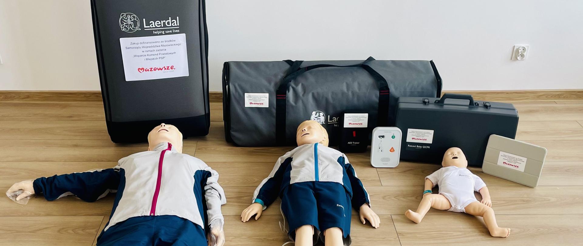 Na zdjęciu znajduje się zestaw fantomów do nauki resuscytacji krążeniowo-oddechowej (RKO) składający się z trzech manekinów: osoby dorosłej, dziecka i niemowlęcia. Manekiny ubrane są w szkoleniowe stroje – dorosły i dziecko mają granatowe spodnie i jasne kurtki, a niemowlę białe body. Za nimi ustawione są torby transportowe i walizki z logo firmy Laerdal oraz oznaczeniami projektu „Mazowsze”, zawierające akcesoria szkoleniowe, w tym symulatory defibrylatorów AED. Całość ustawiona jest na drewnianej podłodze, przy jasnej ścianie.