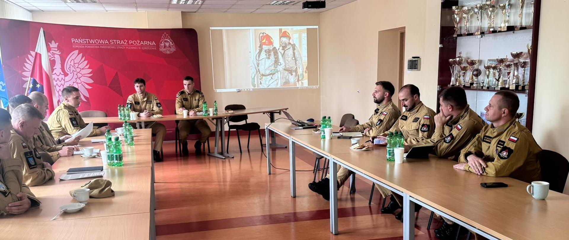Zdjęcie obrazuje podsumowanie ćwiczeń powiatowych pod kryptonimem "Grot 2026". W podsumowaniu ćwiczeń uczestniczyli- naczelnik Wydziału Planowania Operacyjnego Komendy Wojewódzkiej PSP w Kielcach mł. bryg. Kamil Ciepielewski oraz zastępca komendanta powiatowego PSP w Jędrzejowie st. kpt. Tomasz Kwiecień oraz rozjemcy, którzy oceniali ćwiczenia.