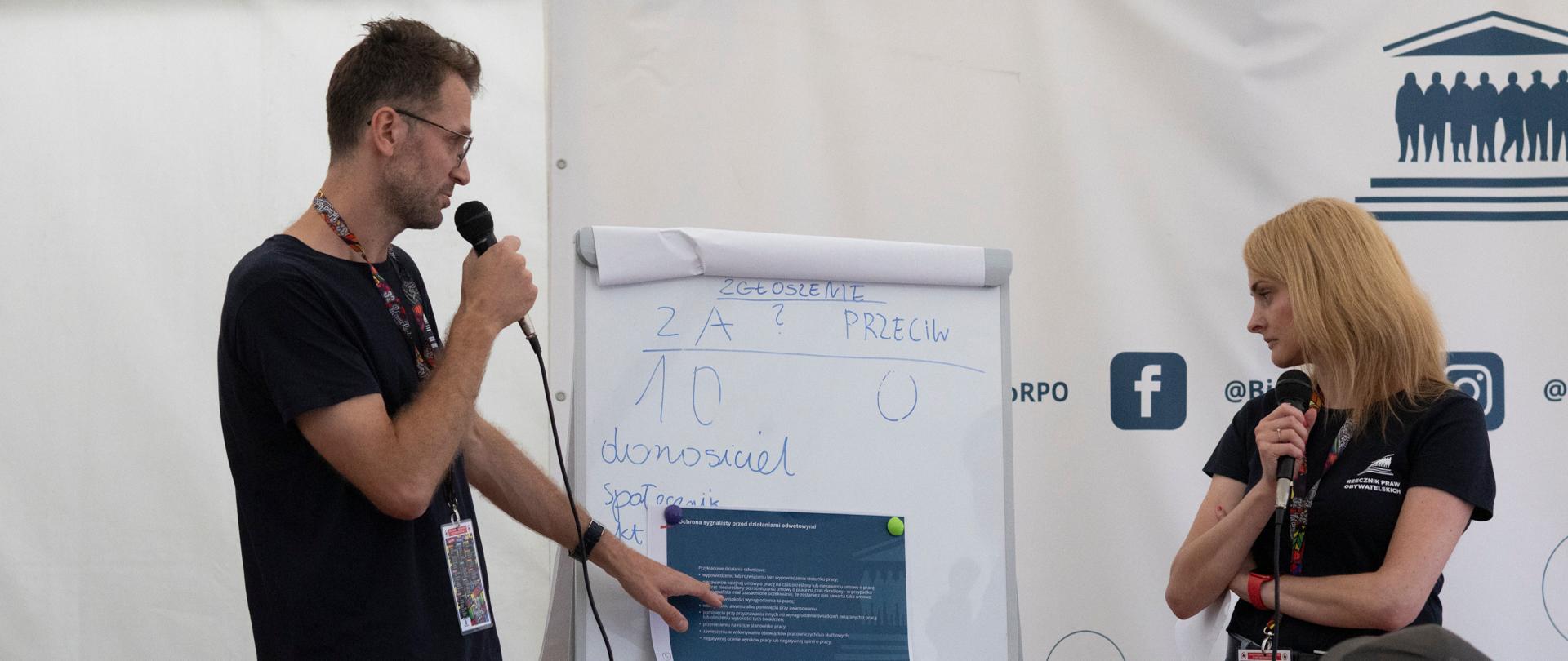 Dwie osoby prowadzą warsztaty w dużym namiocie. Obok nich stoi tablica, na której znajdują się materiały edukacyjne.