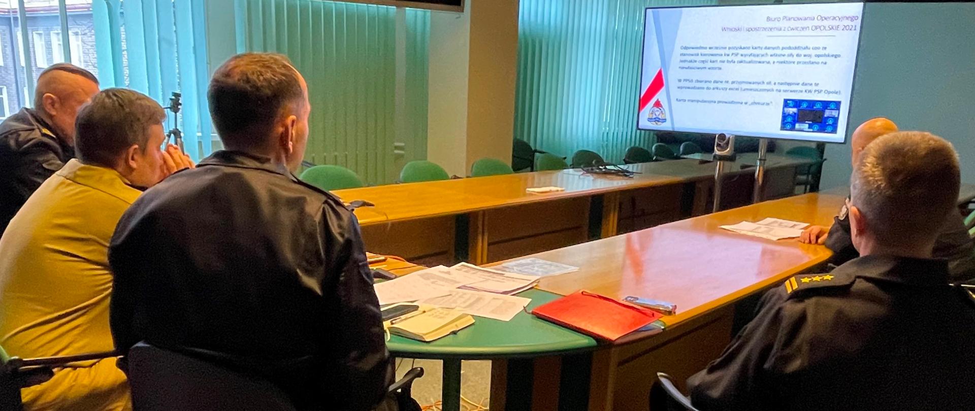 5 funkcjonariuszy siedzi przy stole (widok z tyłu) , przed nimi w głębi sali ekran, na którym wyświetla się slajd z napisami