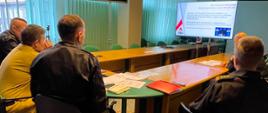 5 funkcjonariuszy siedzi przy stole (widok z tyłu) , przed nimi w głębi sali ekran, na którym wyświetla się slajd z napisami