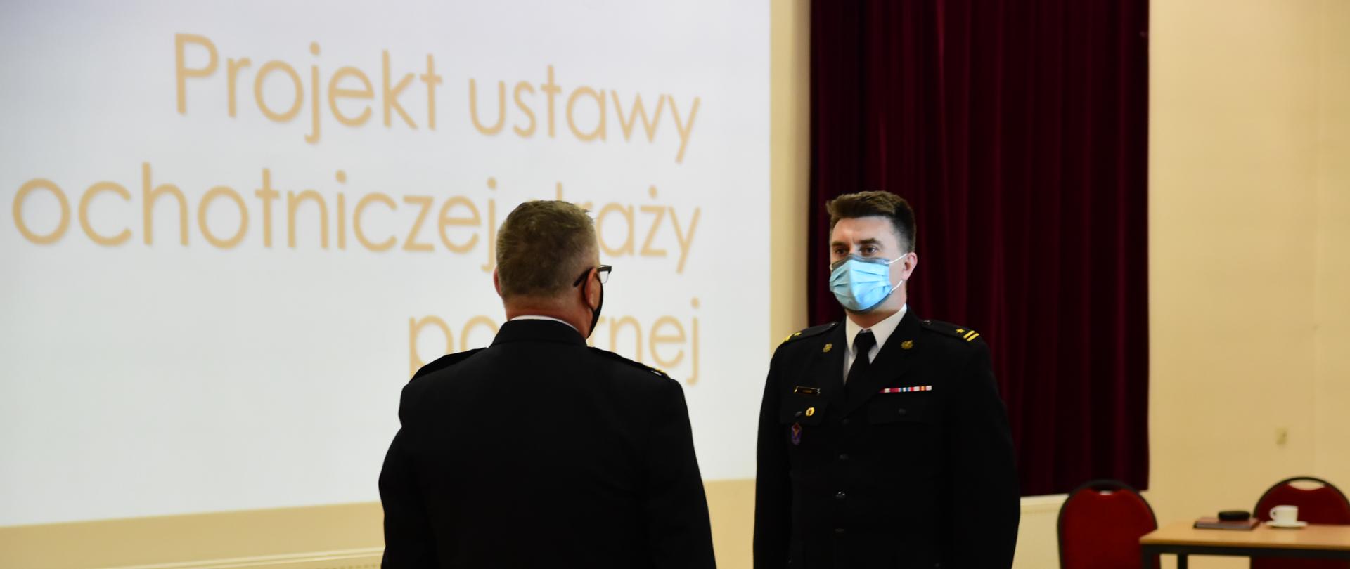 Kujawsko-Pomorski Komendant Wojewódzki Państwowej Straży Pożarnej st.bryg. Jacek Kaczmarek przyjmuje meldunek o gotowości do spotkania w sprawie projektu ustawy o ochotniczej straży pożarnej. Meldunek składa Zastępca Komendanta Powiatowego PSP w Tucholi mł.bryg. Arkadiusz Kroll. W tle widać slajd prezentacji dotyczącej projektu ustawy. Spotkanie odbywa się na sali widowiskowej Ochotniczej Straży Pożarnej w Bysławiu.