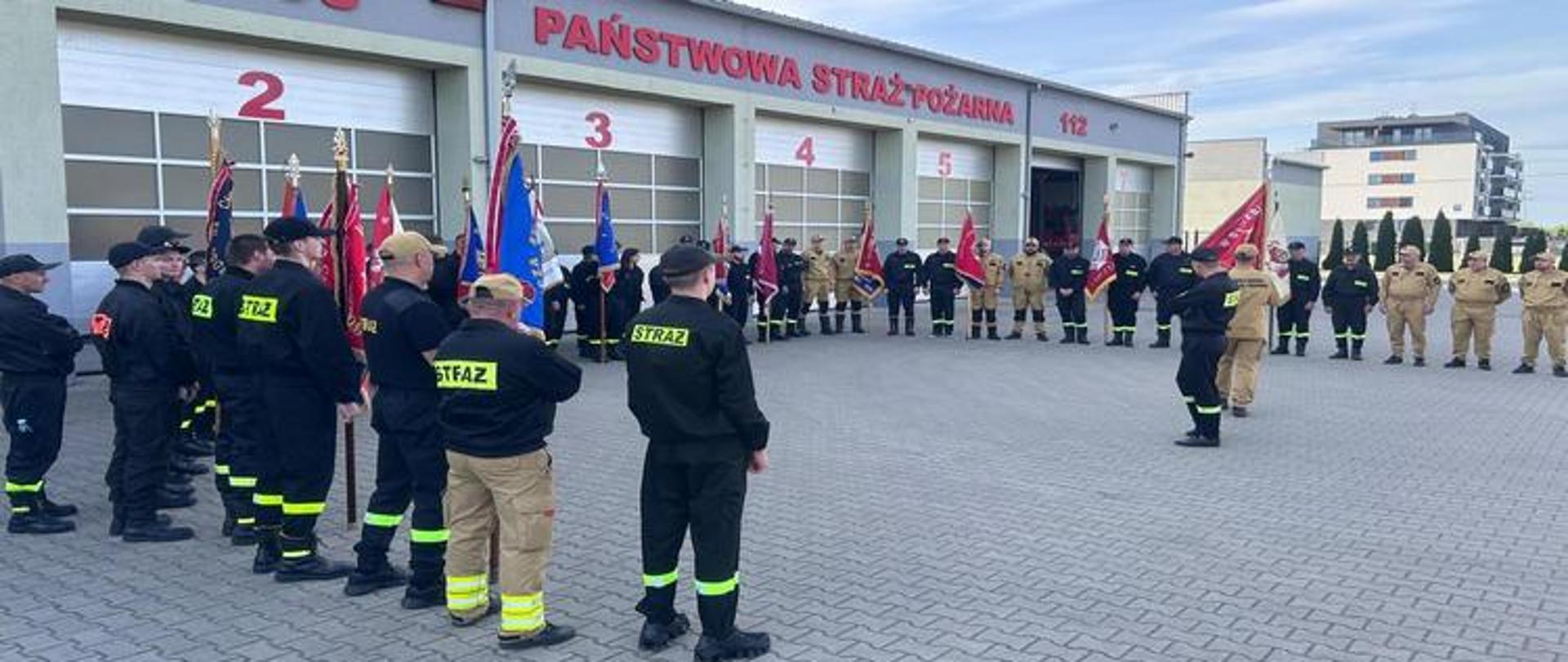 Zdjęcie przedstawia dużą grupę strażaków zebranych na wybrukowanym placu przed budynkiem Państwowej Straży Pożarnej w Sokołowie Podlaskim, co wskazuje napis "PAŃSTWOWA STRAŻ POŻARNA" na froncie budynku. Uroczystość ma charakter oficjalny, na co wskazują liczne poczty sztandarowe z flagami i sztandarami różnych jednostek, widoczne w centralnej części kadru, pomiędzy dwoma głównymi grupami strażaków.
Po lewej stronie znajduje się szereg strażaków ubranych w ciemne (czarne) mundury, a także w jasnobeżowe mundury bojowe, stojących na baczność. Po prawej stronie placu stoi kolejna grupa strażaków w podobnych mundurach, tworząc półokrąg lub otwartą formację. Wśród nich widać także strażaków w galowych mundurach.