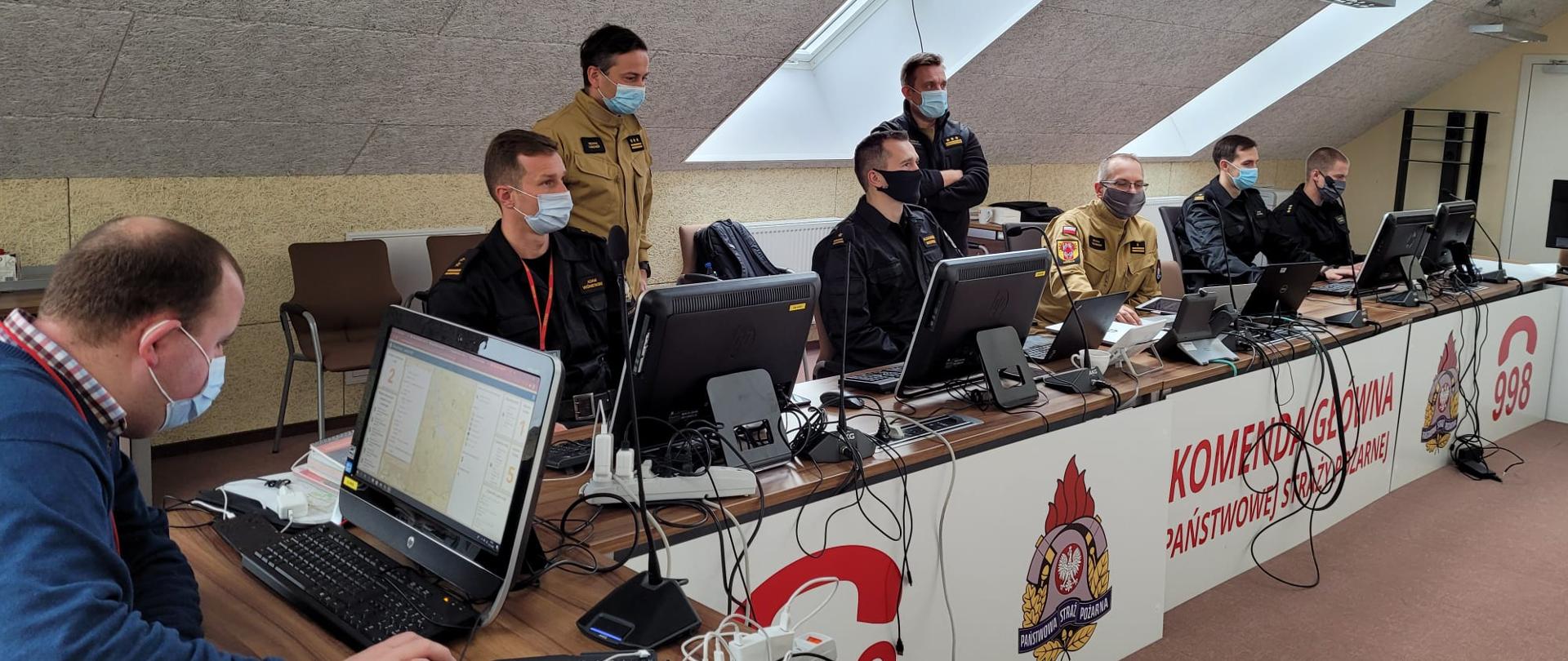 Funkcjonariusze i jeden mężczyzna cywilny (pierwszy od lewej) siedzą przy biurkach podczas ćwiczeń sztabowych, dwóch funkcjonariuszy stoi