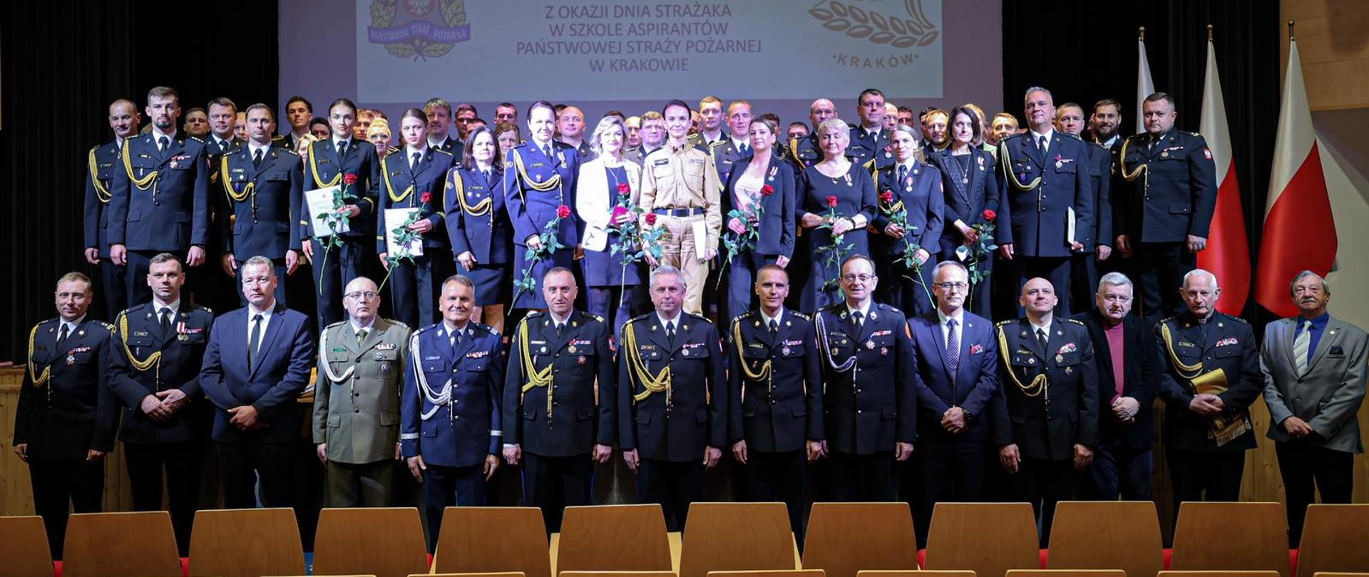 Pamiątkowe zdjęcie uczestników uroczystości
Grupa strażaków w galowych mundurach oraz pracowników cywilnych pozuje na scenie. W tle widoczny jest ekran z napisem „UROCZYSTA ZBIÓRKA Z OCCAZJI DNIA STRAŻAKA W SZKOLE ASPIRANTÓW PAŃSTWOWEJ STRAŻY POŻARNEJ W KRAKOWIE”.
