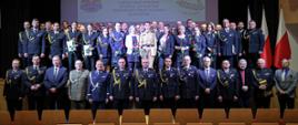 Pamiątkowe zdjęcie uczestników uroczystości
Grupa strażaków w galowych mundurach oraz pracowników cywilnych pozuje na scenie. W tle widoczny jest ekran z napisem „UROCZYSTA ZBIÓRKA Z OCCAZJI DNIA STRAŻAKA W SZKOLE ASPIRANTÓW PAŃSTWOWEJ STRAŻY POŻARNEJ W KRAKOWIE”.

