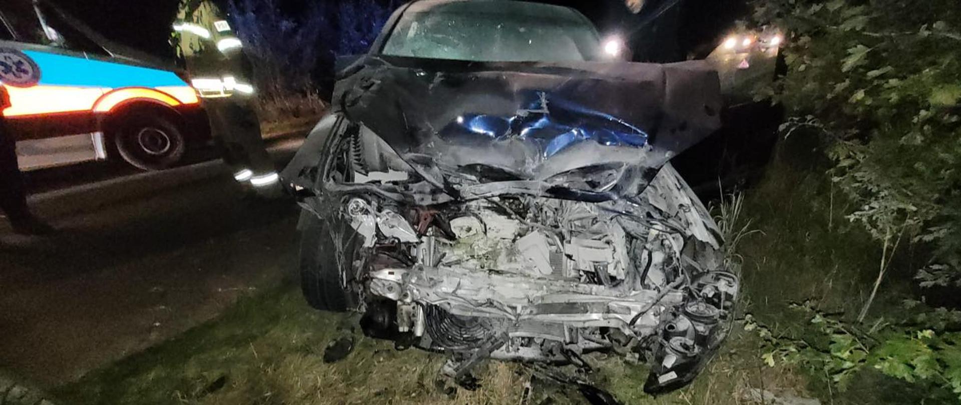 Samochód osobowy z poważnie uszkodzonym przodem po wypadku drogowym nocą. Pojazd stoi na poboczu w trawie, z rozbitym silnikiem i zgniecioną maską. W tle widoczni strażacy oraz karetka pogotowia z włączonymi światłami.