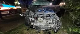 Samochód osobowy z poważnie uszkodzonym przodem po wypadku drogowym nocą. Pojazd stoi na poboczu w trawie, z rozbitym silnikiem i zgniecioną maską. W tle widoczni strażacy oraz karetka pogotowia z włączonymi światłami.