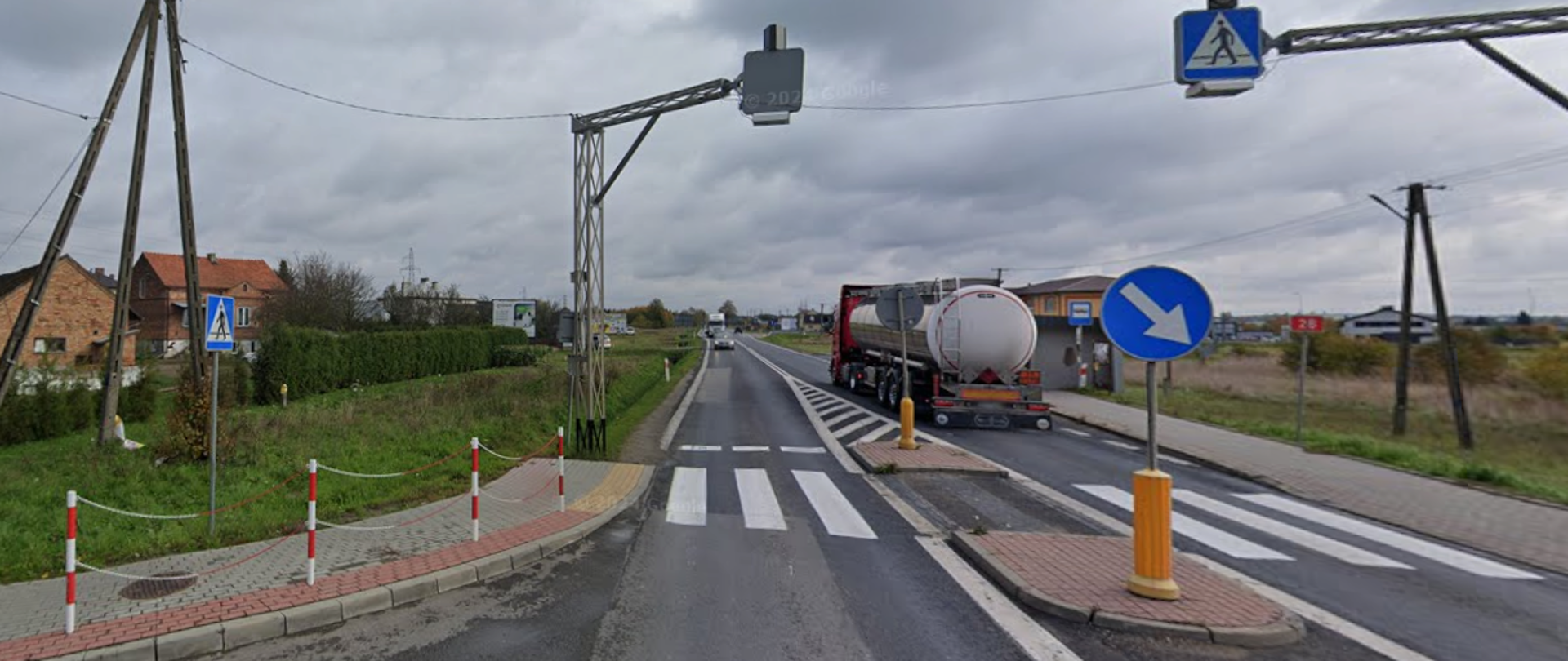 Widok na dwupasmową drogę przebiegającą przez teren podmiejski. Na pierwszym planie znajduje się oznakowane przejście dla pieszych z wyspą dzielącą jezdnię i niebieskimi znakami przejścia. Prawym pasem jedzie duża ciężarówka-cysterna. Po bokach drogi widać chodniki, słupy energetyczne, pojedyncze domy oraz otwarte tereny porośnięte trawą. Nad jezdnią zamontowano konstrukcję z urządzeniem monitorującym ruch. Niebo jest całkowicie zachmurzone, co nadaje scenerii szary, pochmurny charakter.